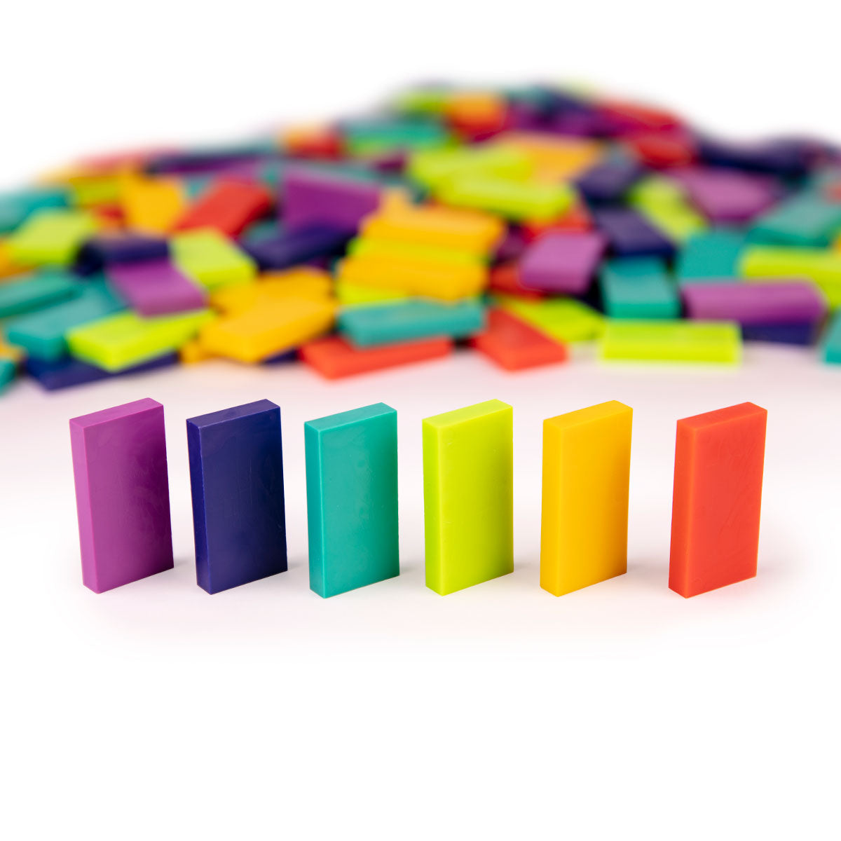 Atwood Toys Bulk Dominoes Inertia Mix, six standing dominoes in raspberry, indigo, teal, sour apple, gold, and lava orange with multi-color domino pile blurred in the background