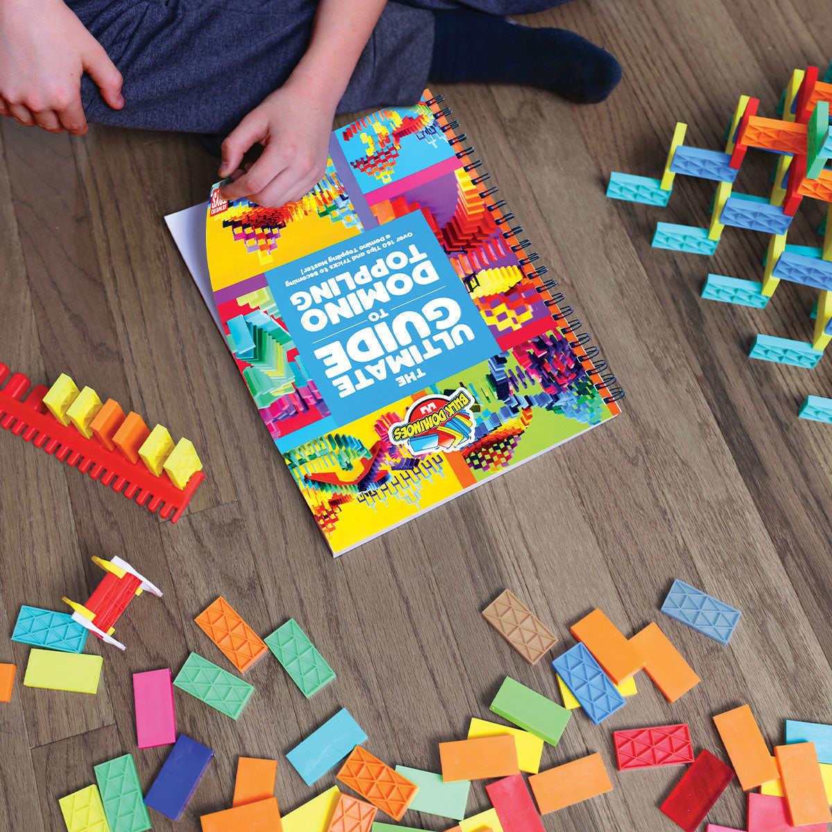 Child reading the Ultimate Guide to Domino Toppling book from the Bulk Dominoes Starter Kit 101, surrounded by rainbow Kinetic Dominoes and 3D structures on a wood floor.