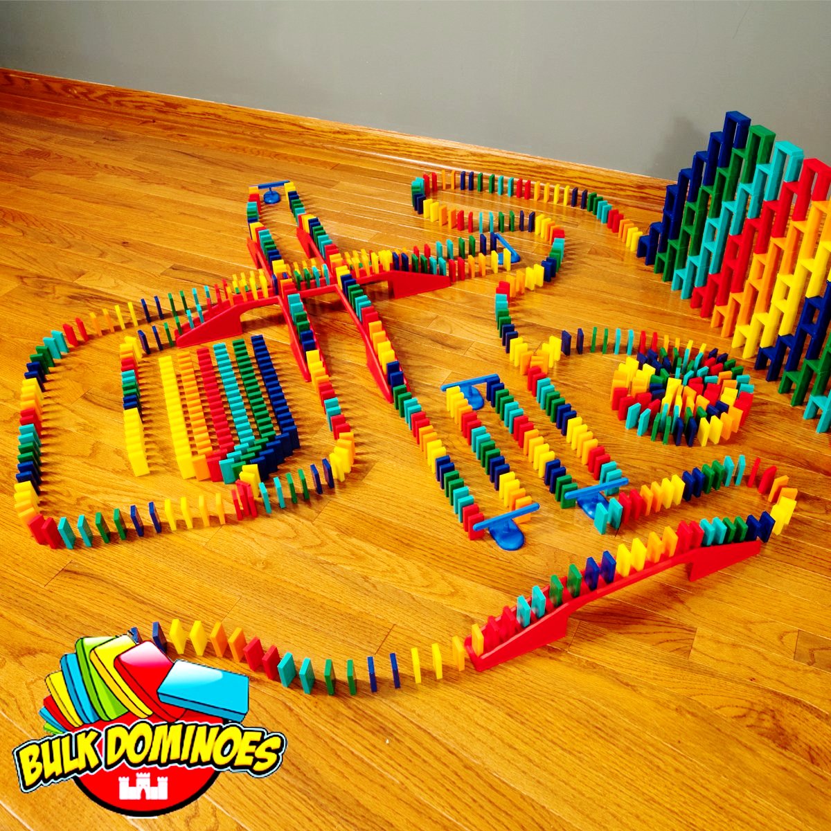 Bulk Dominoes Master Builder Kit setup on hardwood floor shot from above showing colorful Pro Dominoes in winding chain reaction paths, red bridges connecting lanes, and stacked domino walls with Bulk Dominoes logo