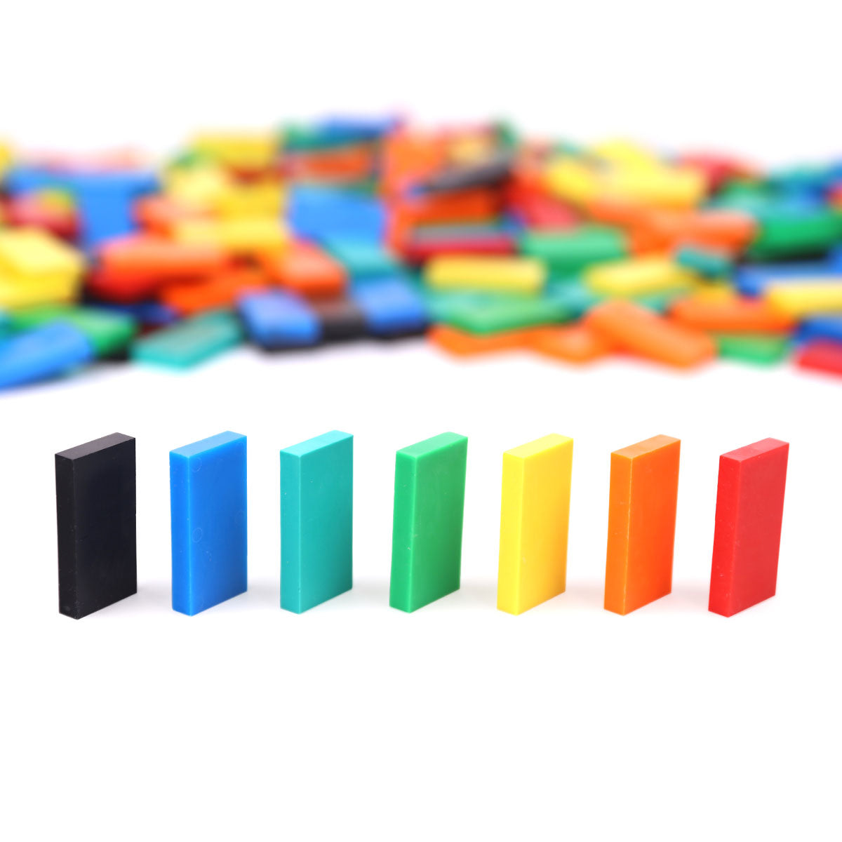 Atwood Toys Bulk Dominoes Classic Mix, seven standing dominoes in black, blue, teal, green, yellow, orange, and red with multi-color domino pile blurred in the background
