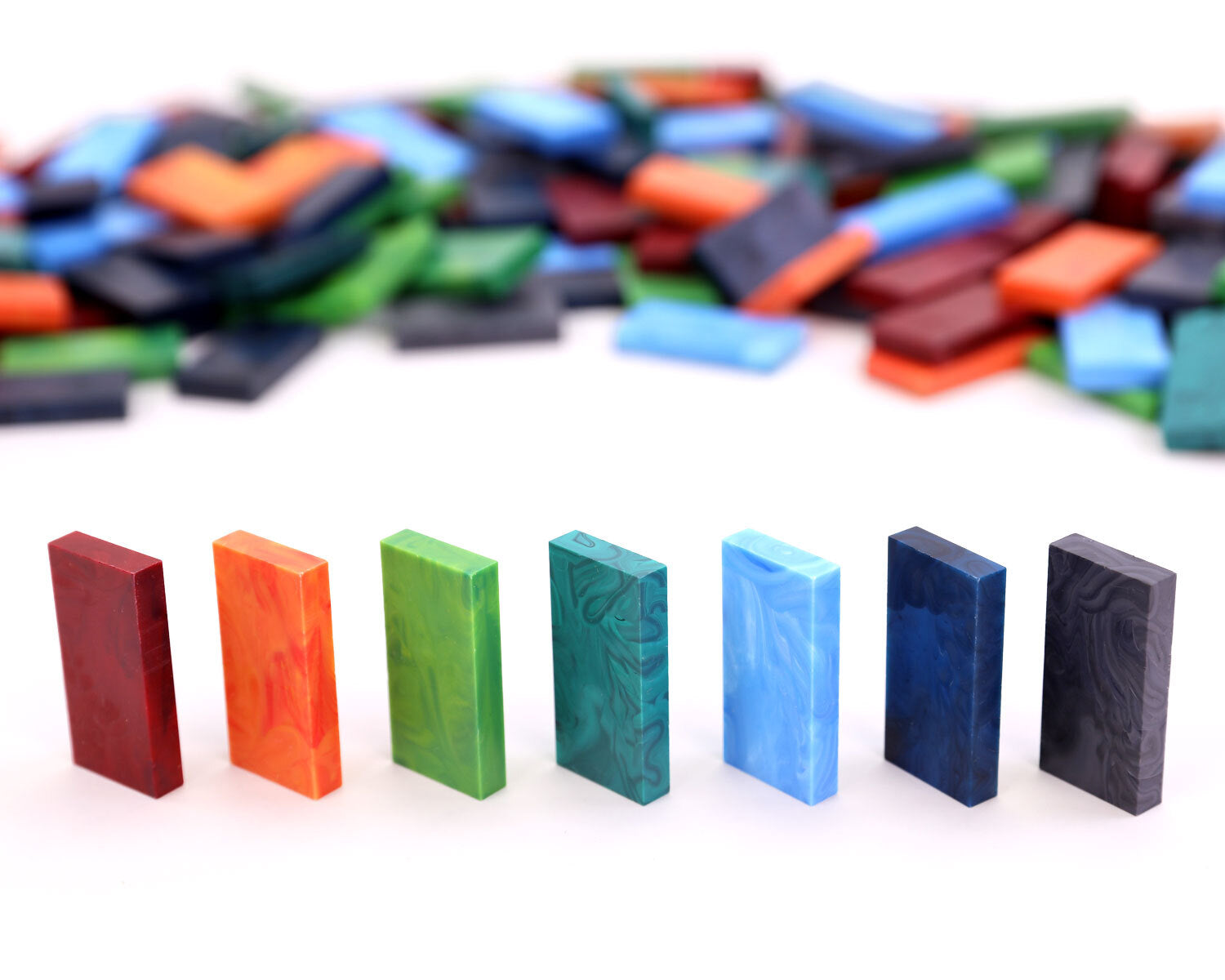Closer view of seven upright Bulk Dominoes Storm Mix Pro Dominoes in marbled red, orange, green, teal, light blue, navy, and black, with a soft-focus pile of matching dominoes in the background.