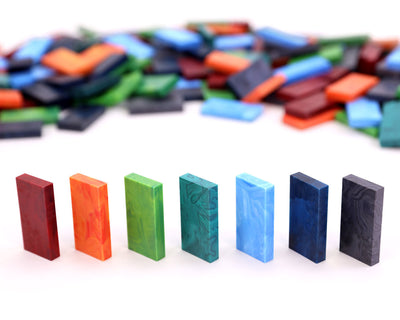 Closer view of seven upright Bulk Dominoes Storm Mix Pro Dominoes in marbled red, orange, green, teal, light blue, navy, and black, with a soft-focus pile of matching dominoes in the background.