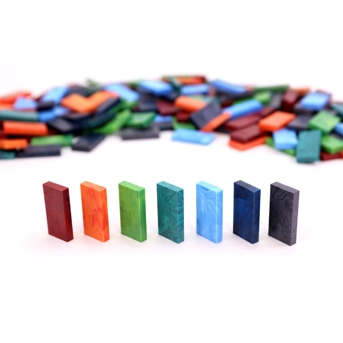 Row of seven Bulk Dominoes Storm Mix Pro Dominoes standing upright in marbled red, orange, green, teal, light blue, navy, and black, with a blurred pile of matching dominoes behind.