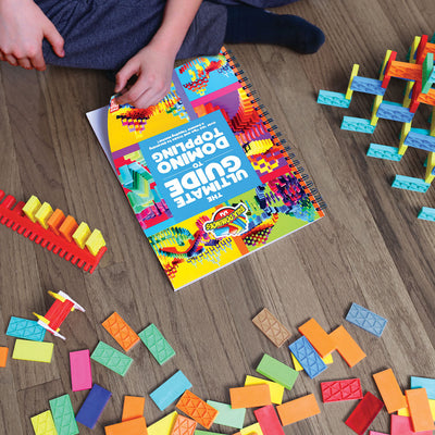 Child reading the Ultimate Guide to Domino Toppling book from the Bulk Dominoes Starter Kit 101, surrounded by rainbow Kinetic Dominoes and 3D structures on a wood floor.
