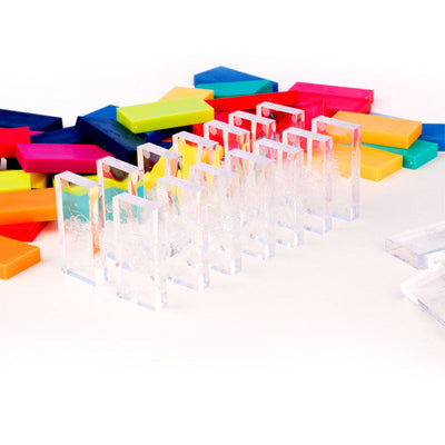 Row of clear transparent Bulk Dominoes Pro Dominoes from Atwood Toys standing in front of a pile of multicolor dominoes including yellow, orange, pink, blue, green, and teal.