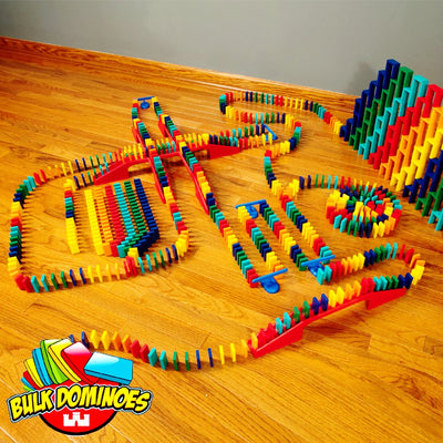 Bulk Dominoes Master Builder Kit setup on hardwood floor shot from above showing colorful Pro Dominoes in winding chain reaction paths, red bridges connecting lanes, and stacked domino walls with Bulk Dominoes logo